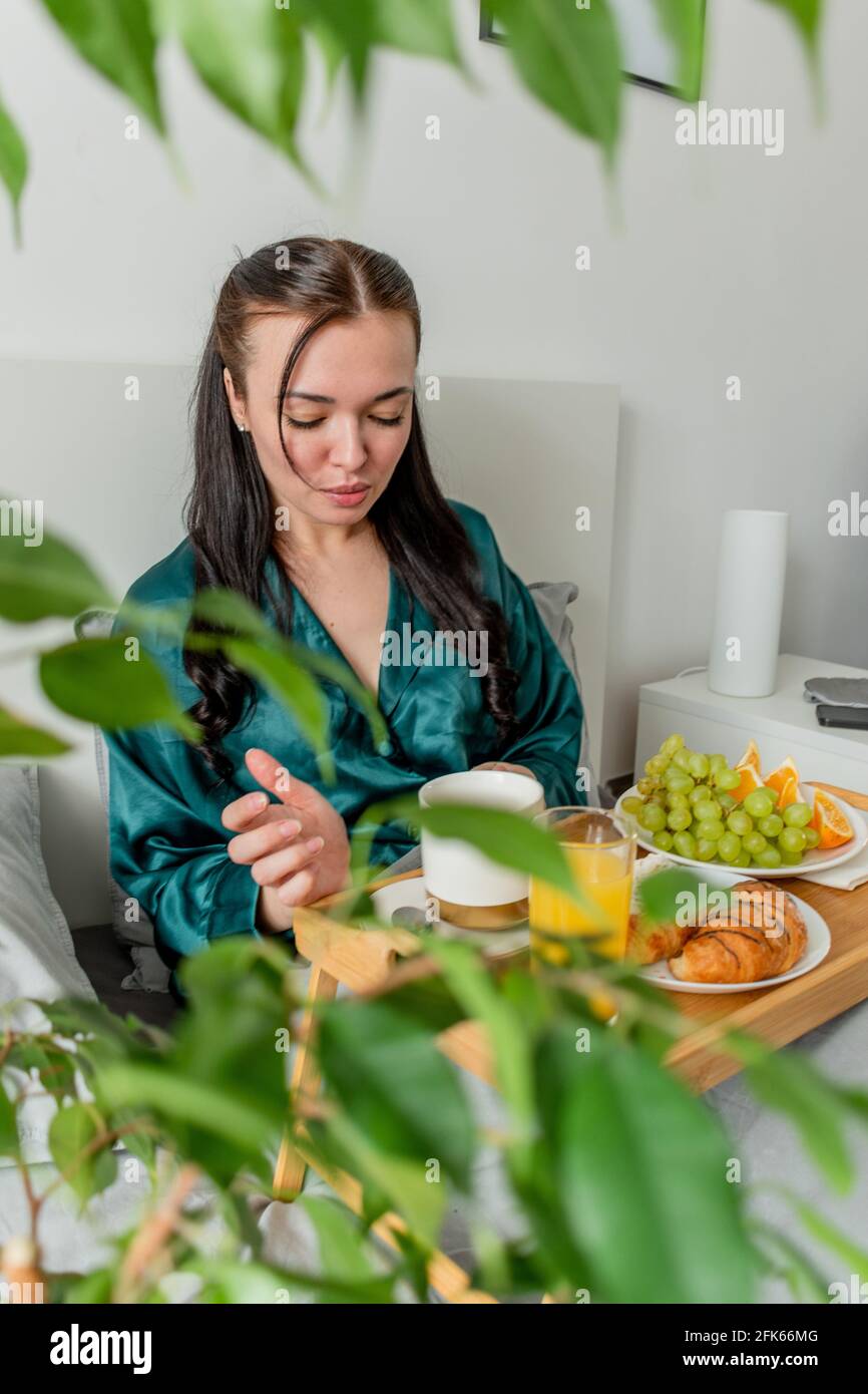 Die junge Frau im Seiden-Pyjama hat ein gesundes Frühstück, während sie im Schlafzimmer auf dem Bett liegt. Konzept für das Wohlbefinden zu Hause. Emotionale Gesundheit einer jungen Frau Stockfoto