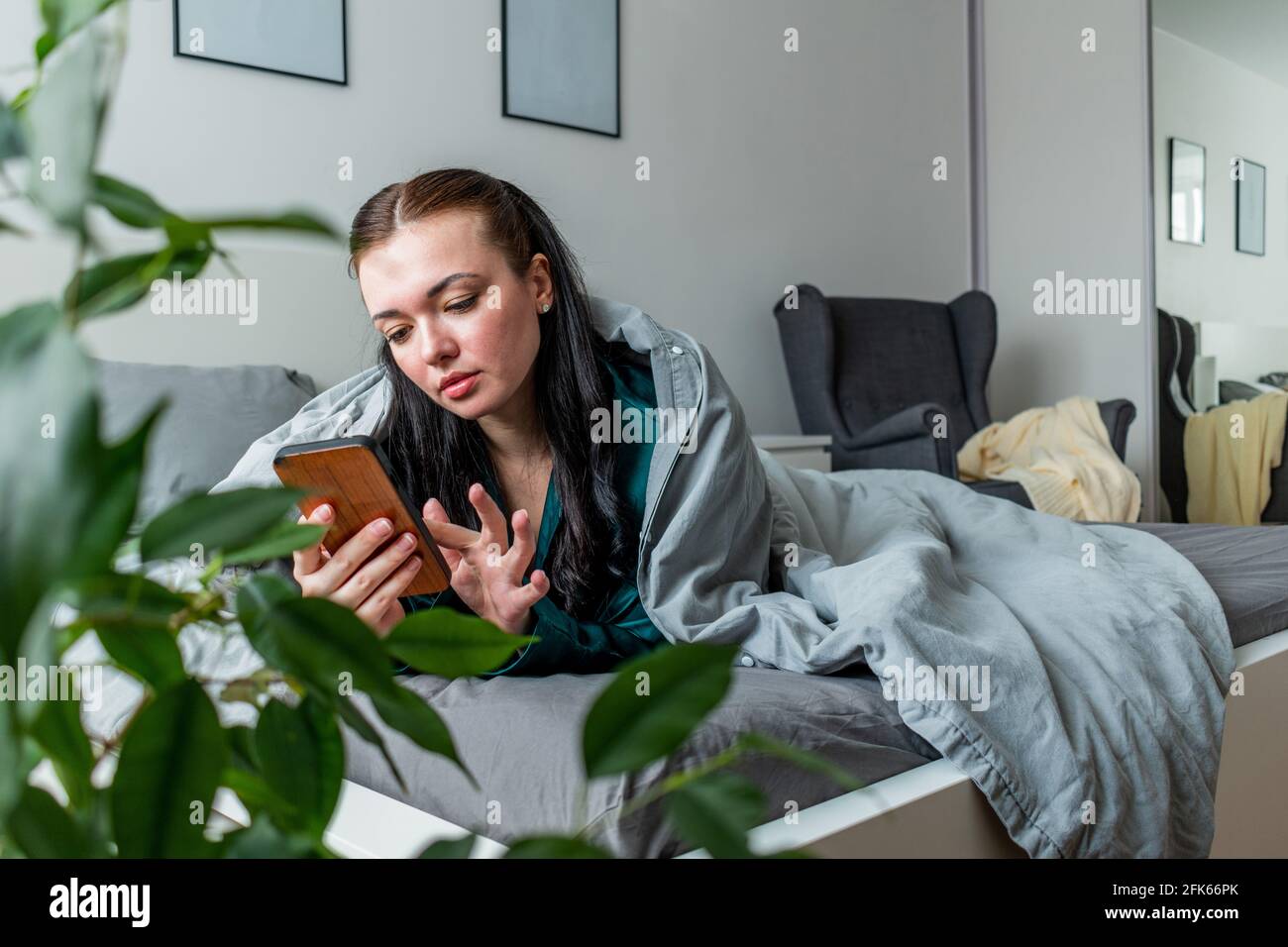 Der Seidenschlafanzug der jungen Frau, der im Bett liegt, hält ihr Handy an den Händen. Arbeiten Sie bequem von zu Hause aus. Wellness-Konzept. Stockfoto