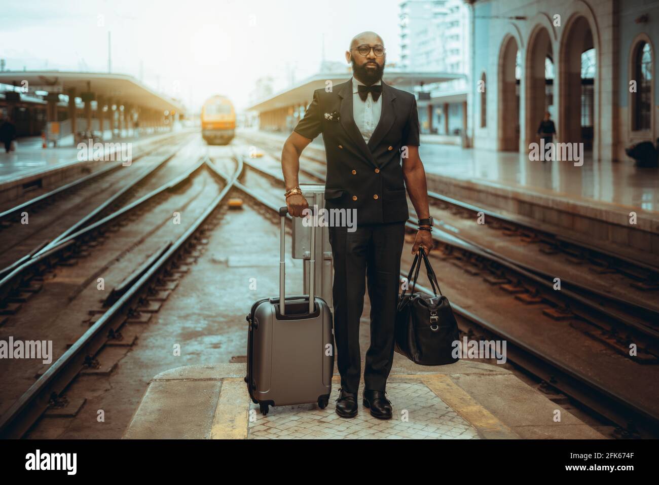 Ein ernsthafter bärtiger, haarloser, gut gekleideter Schwarzmann-Unternehmer in einem Anzug mit Fliege und Gepäck wartet auf dem Die Plattform des Bahnhofs d Stockfoto