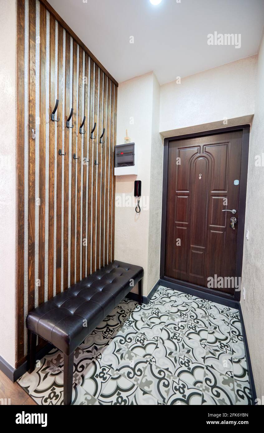 Kompakter Flur mit Holzelementen, auf denen Haken für Oberbekleidung montiert werden. Harmonische Kombination aus braunen Türtönen, gepolsterter Bank, Holzwanddekor und Bodenfliesen. Stockfoto
