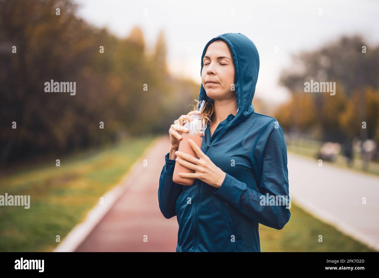 Erwachsene Frau trinkt Wasser, während sie an bewölktem Tag im Herbst im Freien trainiert. Stockfoto