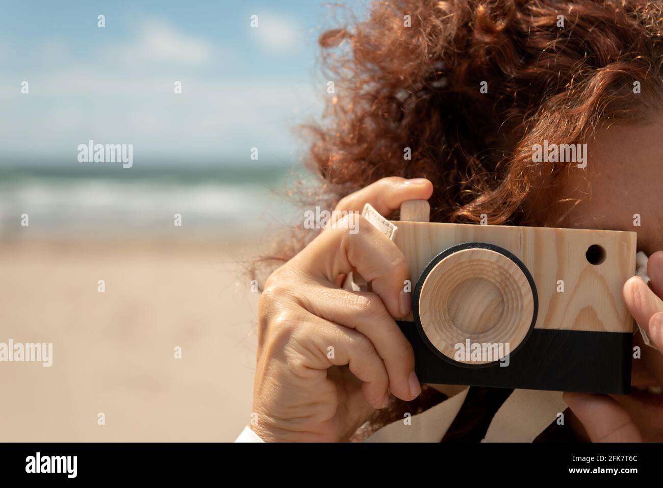 Nahaufnahme eines Porträts einer jungen Frau mit lockigen roten Haaren, die eine Fotokamera mit Holzspielzeug vor dem Hintergrund eines Sandstrandes hält. Witziges Fotografiekonzept Stockfoto