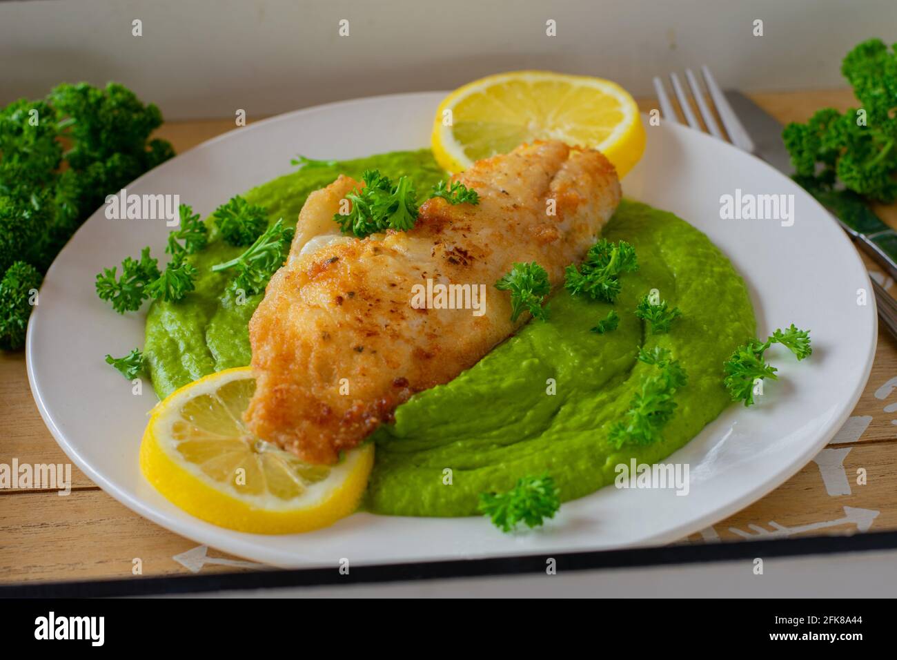 Hausgemachte Mahlzeit mit gebratenem Fisch und grünem Erbsenpüree Serviert mit Zitronenscheiben auf einem weißen Teller mit Messer Und Gabel Stockfoto