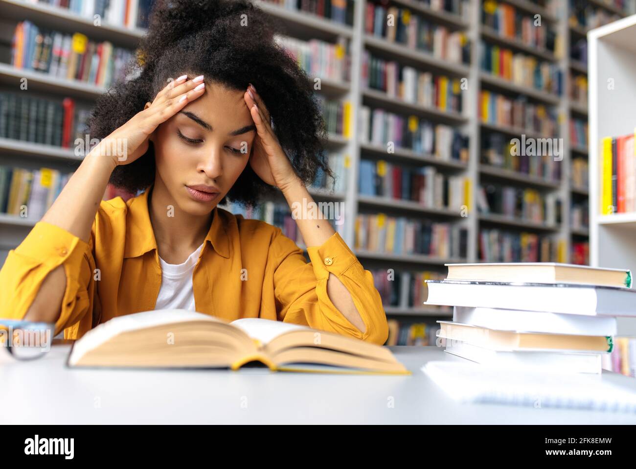 Smart afroamerikanische Studentin, sitzt in stilvoller Kleidung, in der Campus-Bibliothek am Tisch, Buch mit Interesse lesen, Vorbereitung auf Vorlesung oder Prüfung, Wissen und Lernkonzept Stockfoto