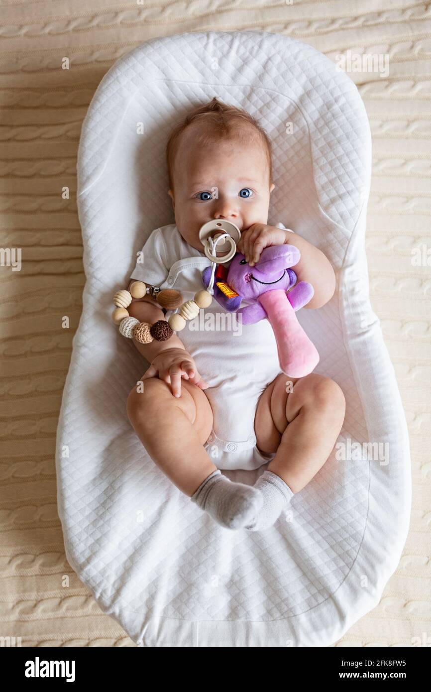 Niedliches blondes Baby, drei Monate alt, das zu Hause im Kokon liegt. Kind hält ausgestopftes Spielzeug, saugt Nippel. Stockfoto