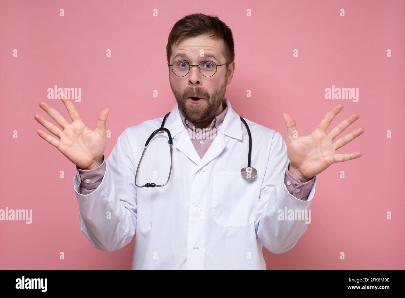 Freudiger Arzt mit einem Stethoskop um den Hals spreizte seine Hände überrascht und blickt bei geöffnetem Mund auf die Kamera. Rosa Hintergrund. Stockfoto