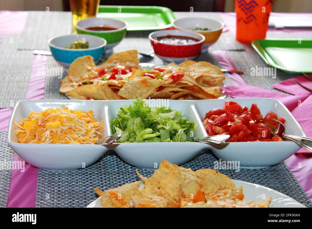 Nahaufnahme eines Tischsets für eine Party, mit Nachos, einem Teller mit Taco-Belägen, einem rosa Tischtuch und Servietten, Platzmatten und Pappteller. Stockfoto