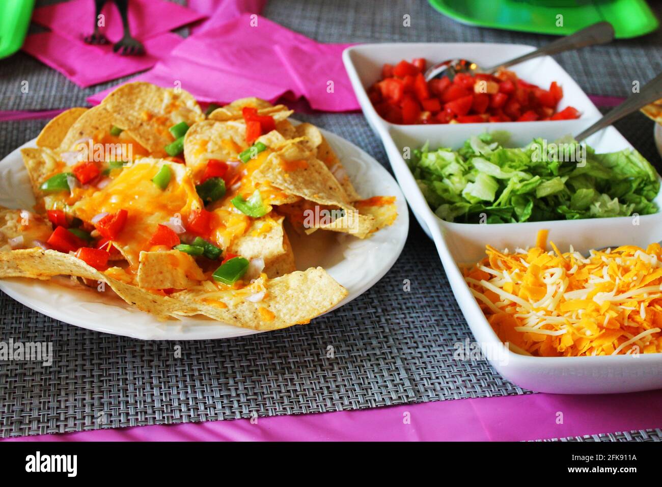 Nahaufnahme eines Tischsets für eine Party, mit Nachos, einem Teller mit Taco-Belägen, einem rosa Tischtuch und Servietten, Platzmatten und Pappteller. Stockfoto
