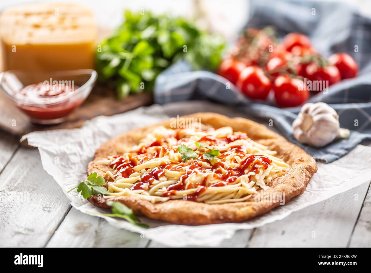 Leckere frittierte Langos auf Backpapier serviert mit viel geriebenem Käse, Ketchup, frischen Kräutern und Tomaten auf einem karierten Geschirrtuch. Stockfoto