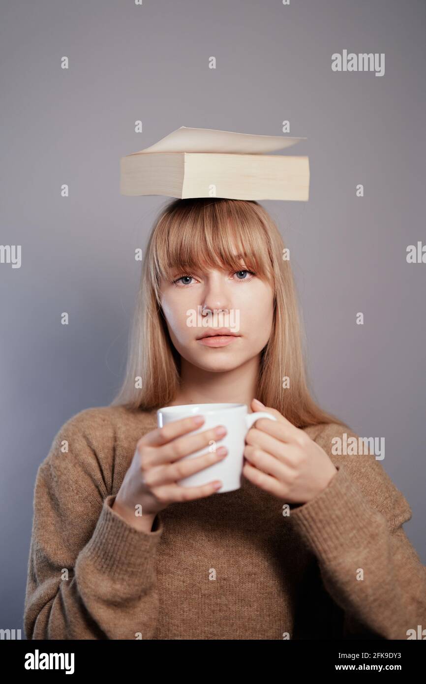 Ernsthaftes Gesichtskonzept. Nerd, Wissens- oder Bildungskonzept. Die attraktive kaukasische Universitätsstudentin trägt eine Brille und einen beigen Pullover mit Buch auf dem Kopf und hält eine Tasse Kaffee oder Tee in den Händen Stockfoto