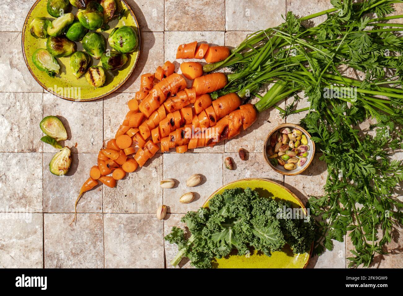 Ein Haufen gegrillter Karottenwurzeln mit grünen Blättern, Rosenkohl, Salatblätter, Nüsse, Aprikosenmarmelade auf Steingeschichten und handgebastelt grüne Teller Draufsicht. Hea Stockfoto
