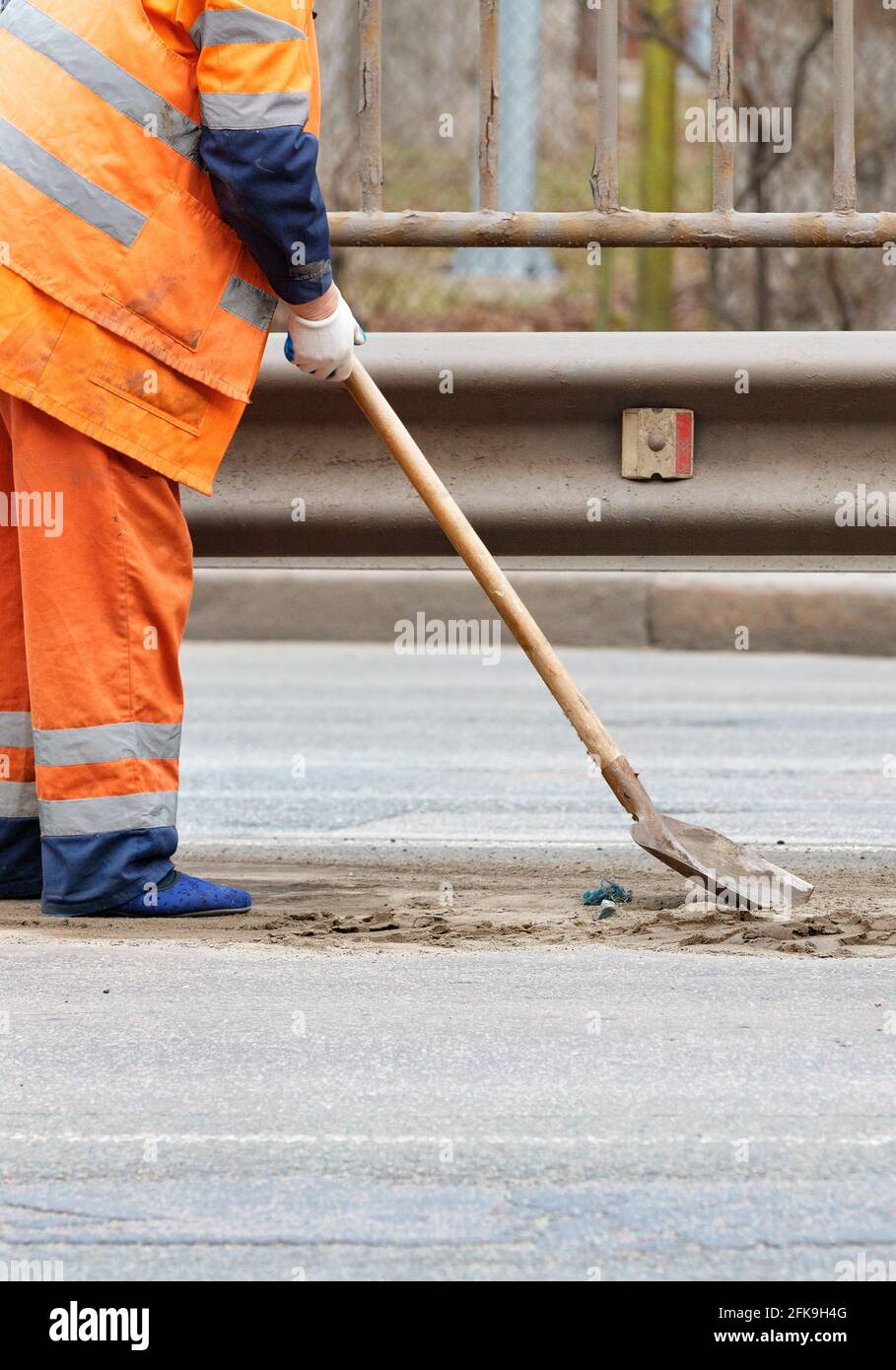 Ein Hausmeister in orangefarbener, reflektierender Uniform schaufelt unter einem Metallstoßfänger Sand von der Straße zwischen den Fahrspuren. Vertikales Bild, Kopierbereich. Stockfoto