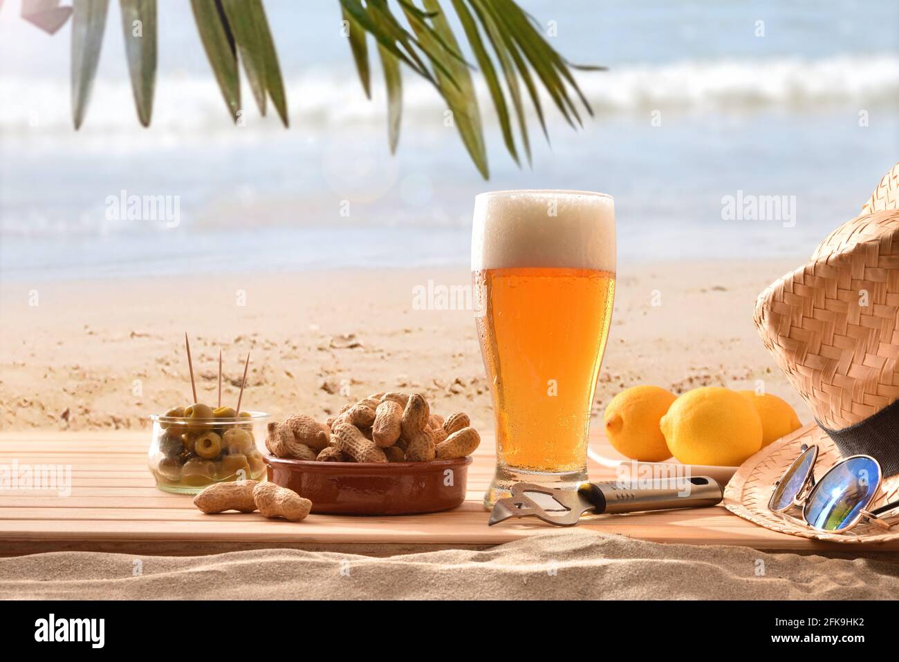 Detail von Glas Bier frisch auf Holz in der Sand zusammen mit einem sukkkkkkulenten Snack an einem Strand mit Palmen an einem heißen Tag Stockfoto