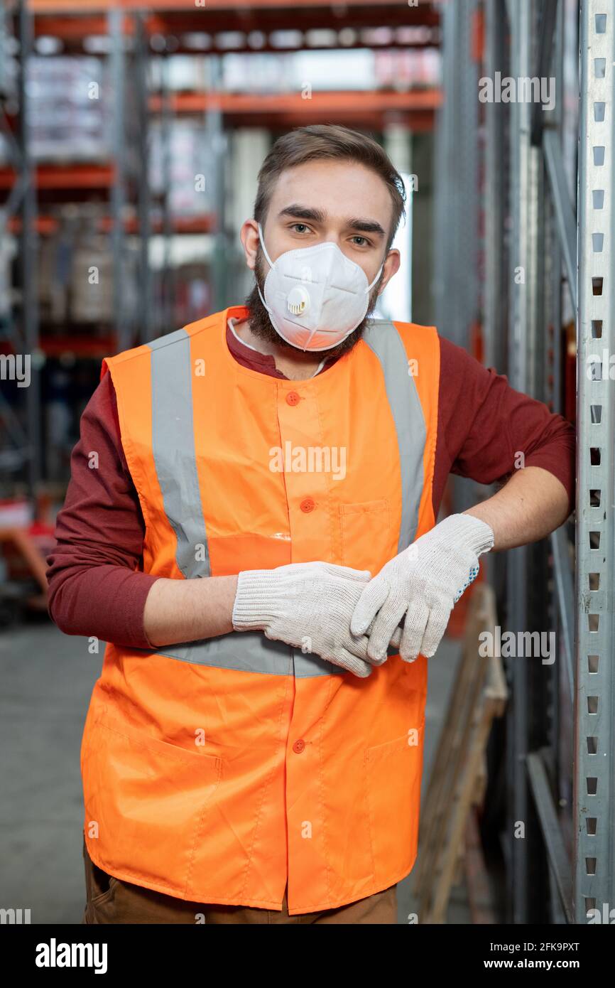 Porträt eines hübschen Schiffsarbeiters in weißer Maske, Handschuhen und reflektierender Weste, die am Metallrahmen im Lager angelehnt ist Stockfoto