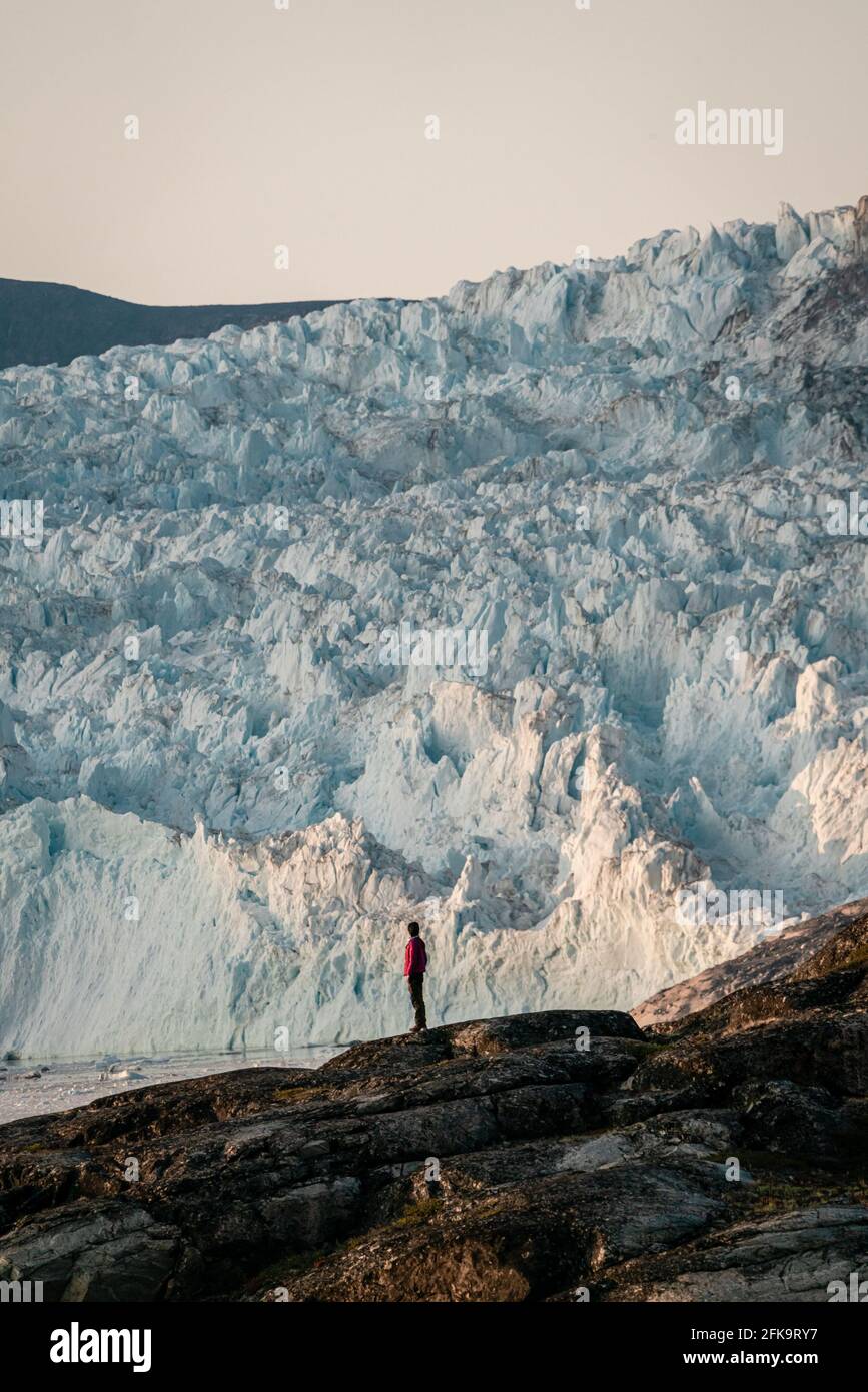 Nicht identifizierte Personen, die vor einer riesigen Gletscherwand aus Eis sitzen. Eqip Sermia-Gletscher der Eqi-Gletscher in Grönland wird als kalbender Gletscher bezeichnet Stockfoto