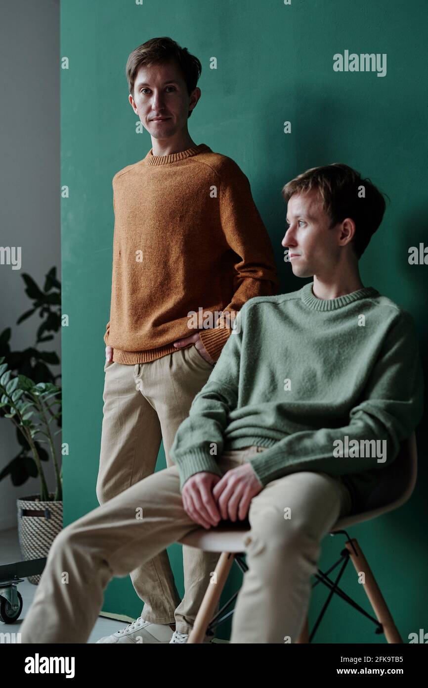 Porträt eines hübschen jungen Mannes in einem Pullover, der die Hände hält In den Taschen und sein Zwillingsbruder sitzt auf Stuhl und Wegschauen Stockfoto