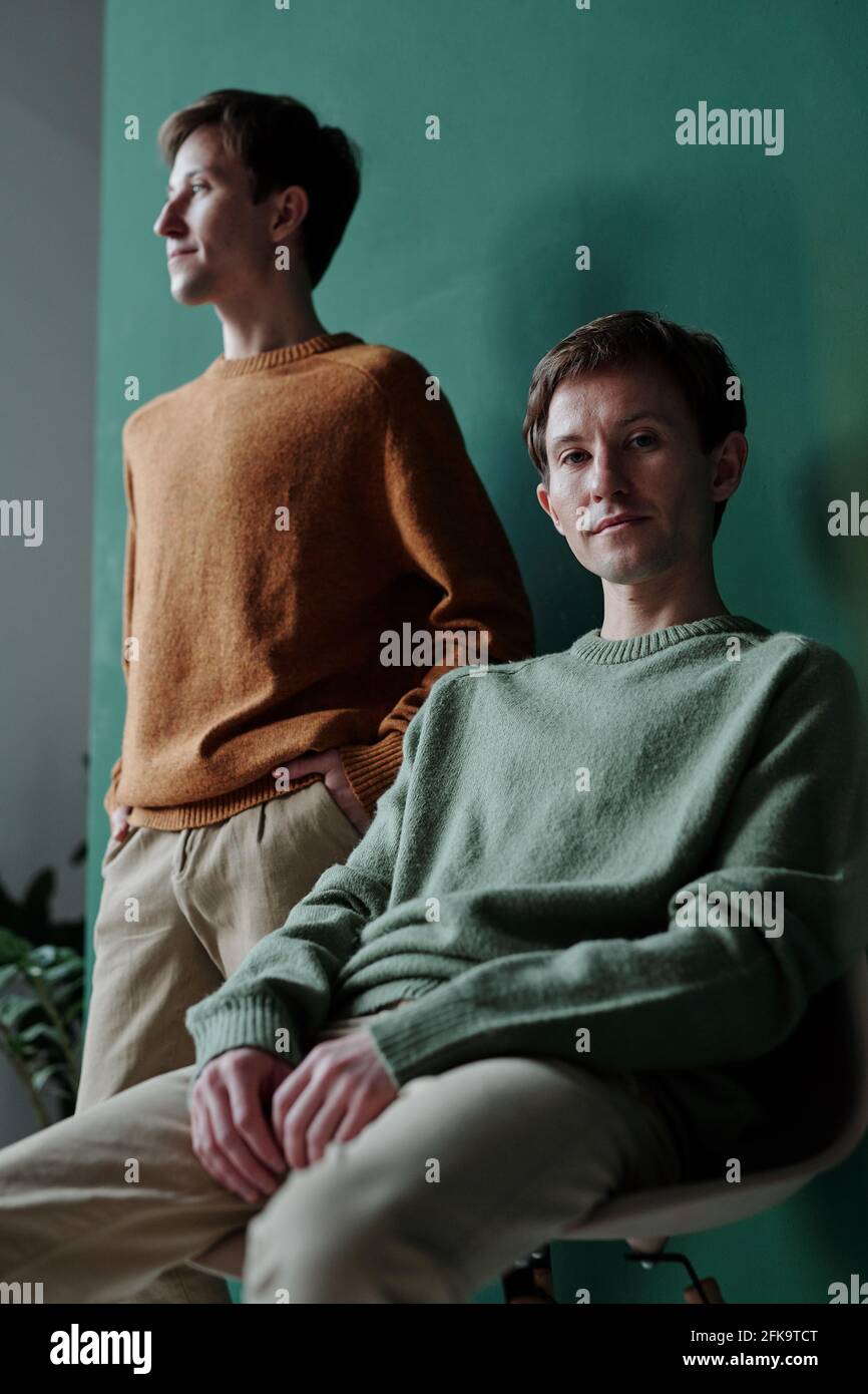 Porträt des Inhalts junger Mann in grünem Pullover sitzend auf Und posiert gegen seinen Zwillingsbruder, der in die Ferne blickt Stockfoto