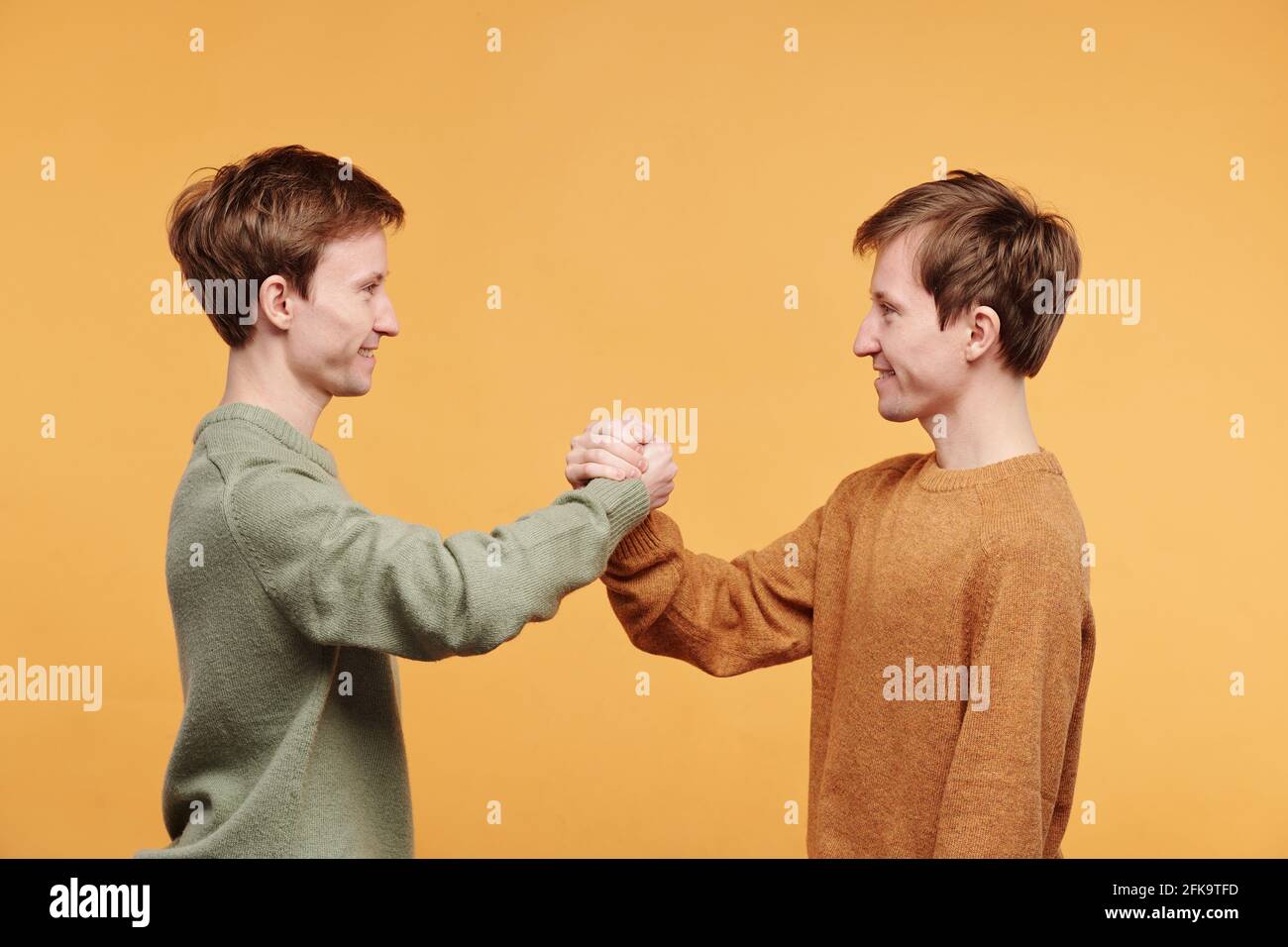 Seitenansicht von lächelnden, freundlichen Zwillingen in Pullovern, die dagegen stehen Orangefarbener Hintergrund und ein fester Händedruck Stockfoto