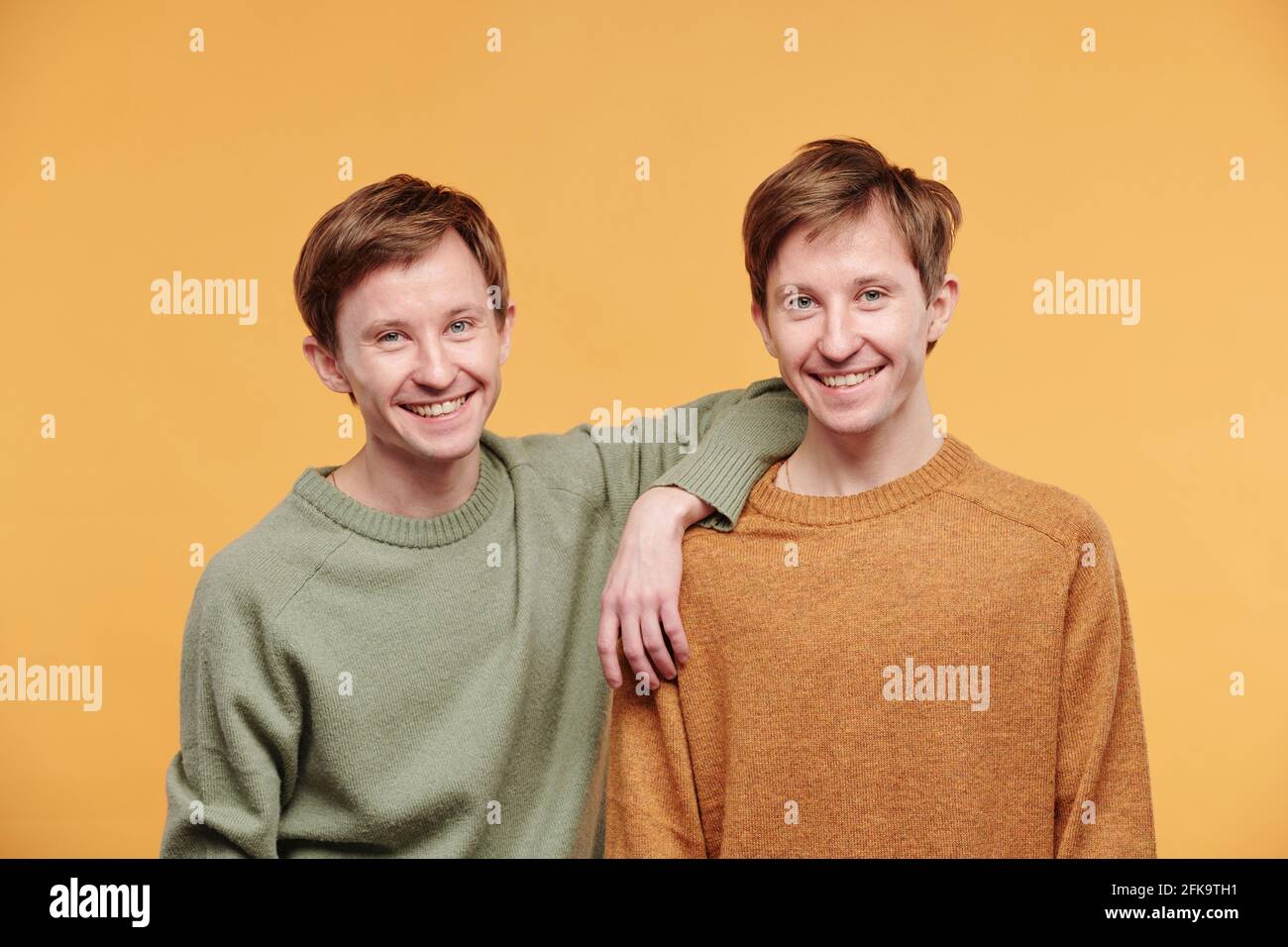Porträt eines glücklichen jungen Mannes in legerem Pullover, der sich anlehnt Schulter des Zwillingsbruders auf orangefarbenem Hintergrund Stockfoto