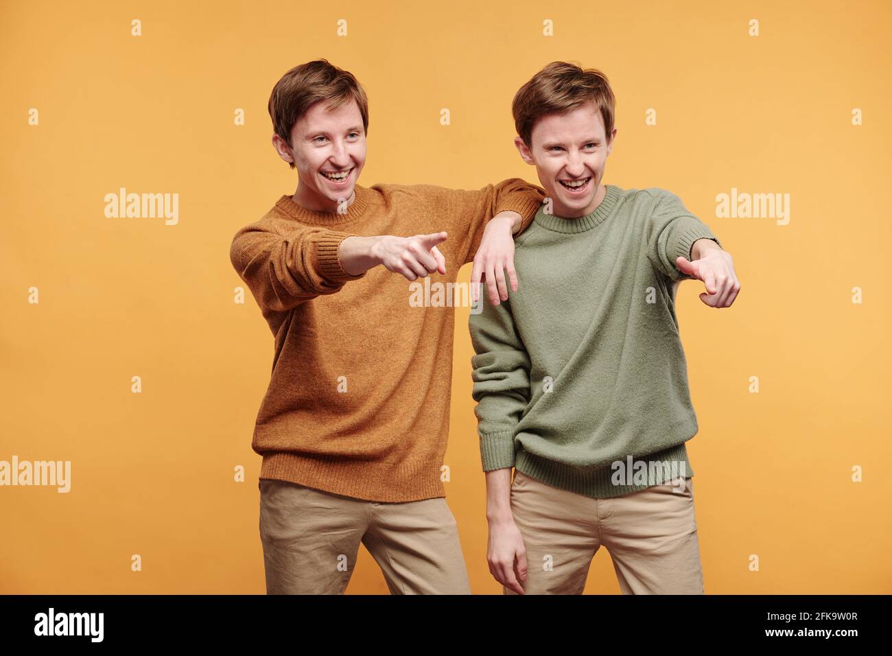 Positive junge Zwillinge in Pullovern, die lachen, während sie auf etwas zeigen Vor orangefarbenem Hintergrund Stockfoto