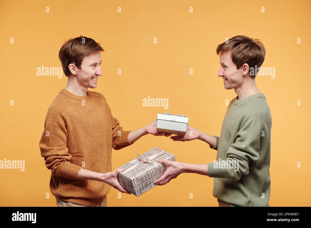 Fröhliche junge Zwillinge in Pullovern stehen vor orangefarbenem Hintergrund und tauschen Sie Geschenke aus, und gratulieren Sie sich gegenseitig zum Geburtstag Stockfoto