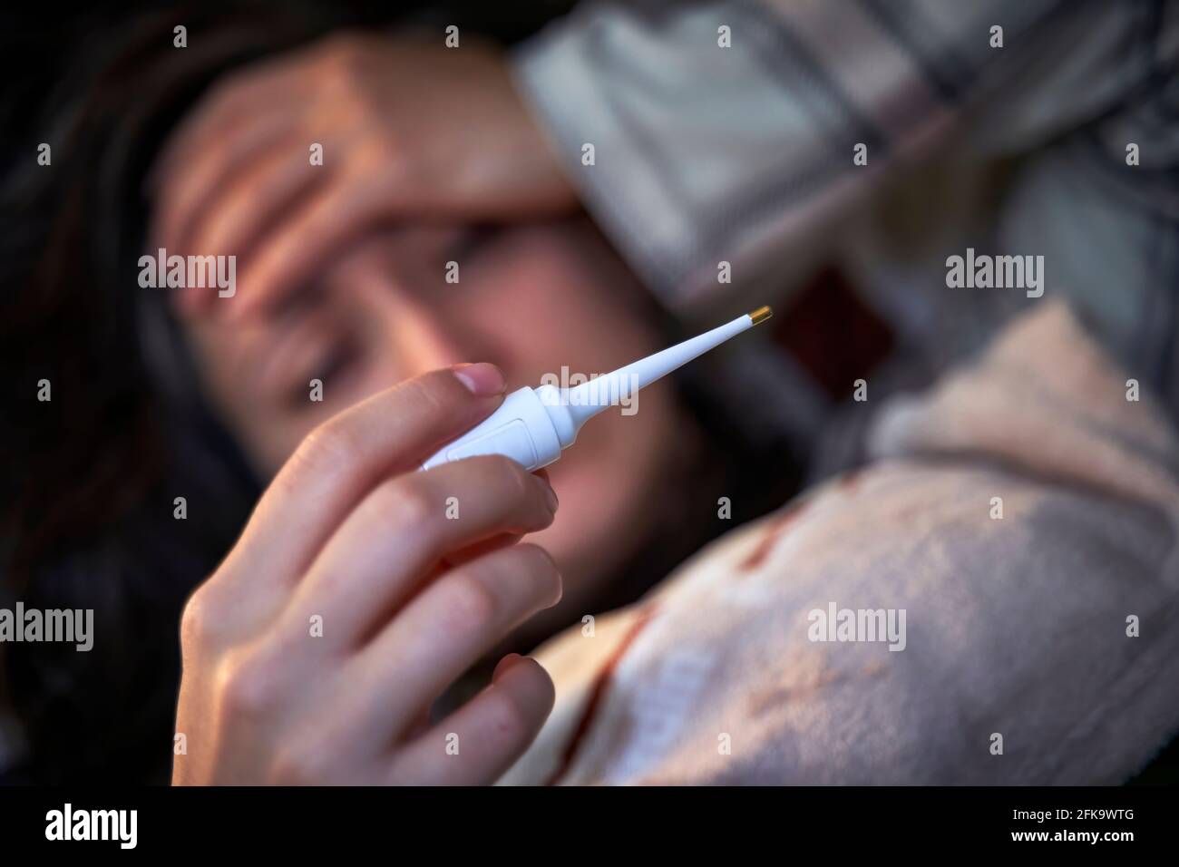 Verschwommene junge Frau, die sich krank fühlt und Fieber hat, und auf das Thermometer schaut, das ihre Stirn berührt. Konzept von Krankheit und Fieber. Stockfoto
