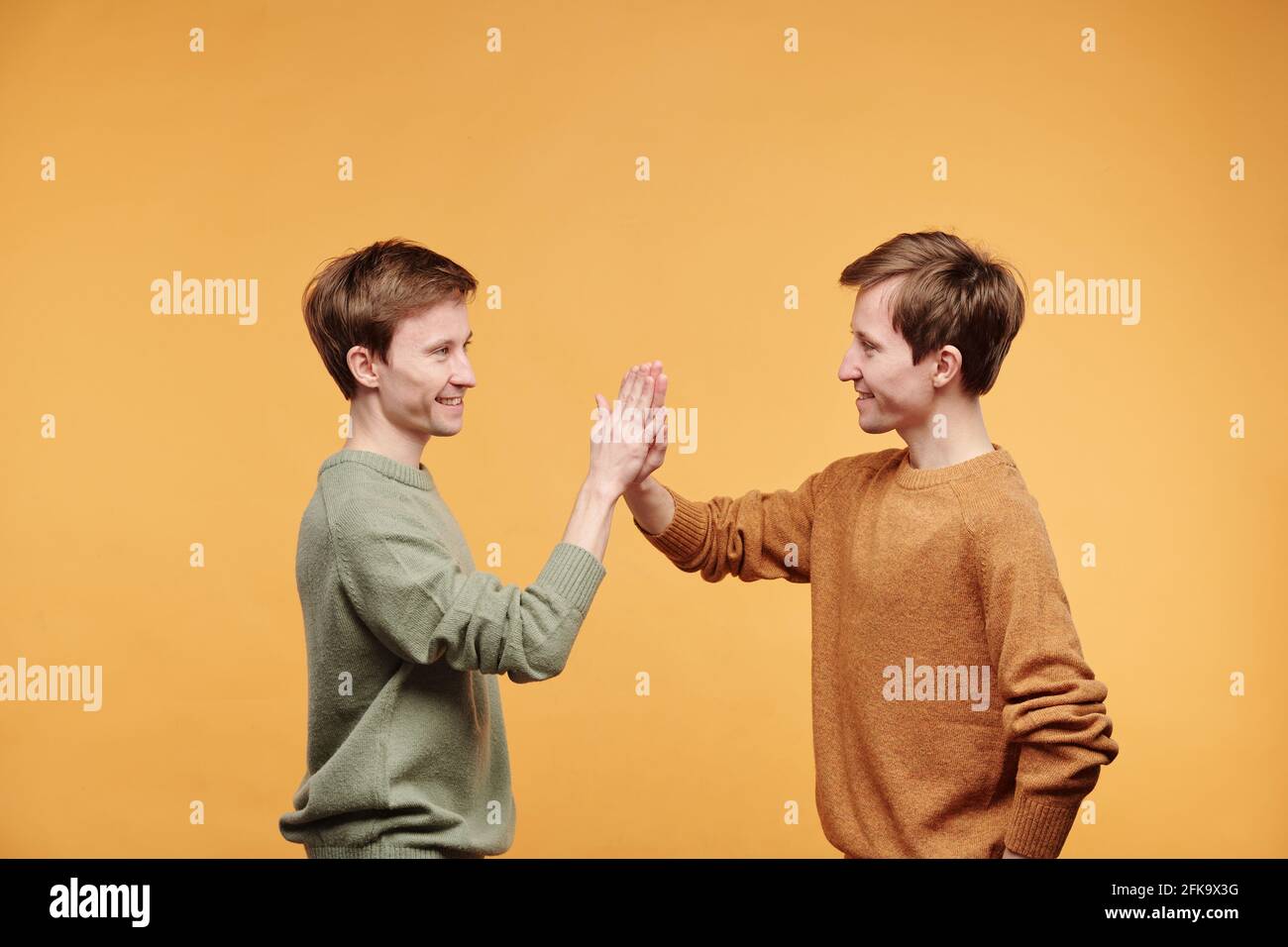 Seitenansicht von gutaussehenden jungen Zwillingen in lässigen Pullovern Vor orangefarbenem Hintergrund stehend und fünf geben Stockfoto