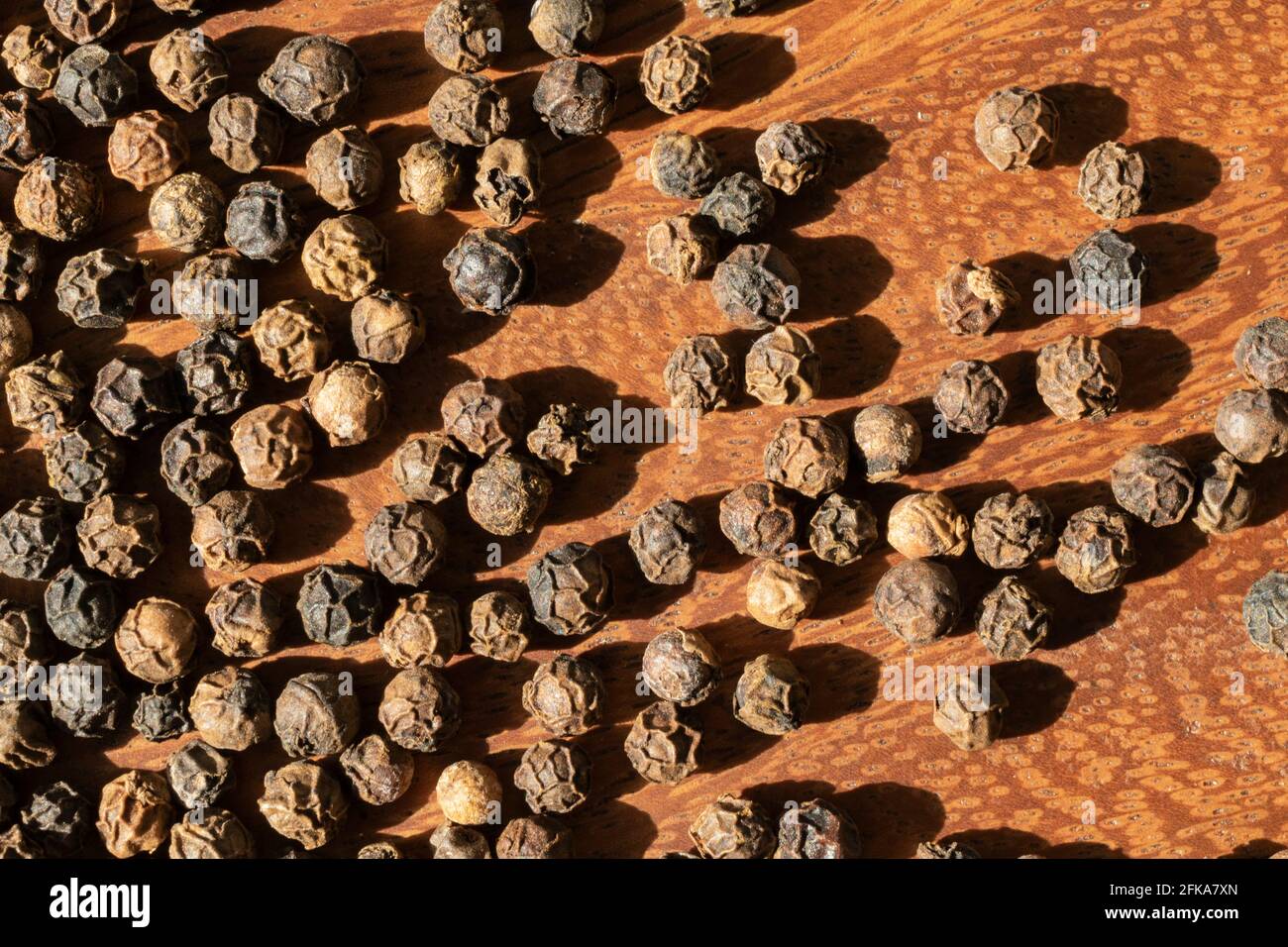 Würzige schwarze Pfefferkörner, Piper nigrum, in der Nachmittagssonne auf einer Holzoberfläche verstreut. Stockfoto