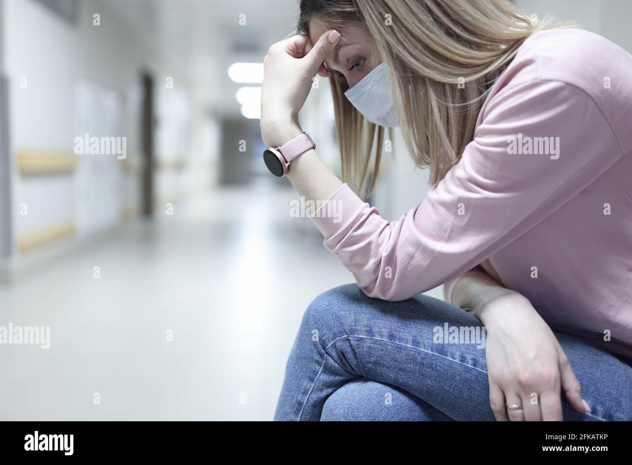 Traurige Frau in medizinischer Schutzmaske sitzt im Flur von Klinik Stockfoto