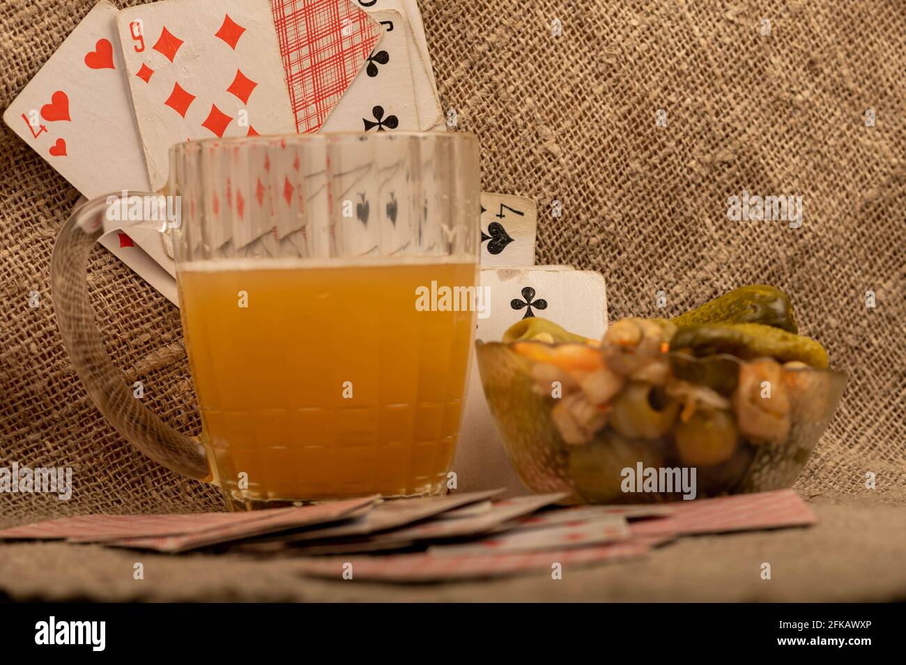 Spielkarten, ein Becher mit leichtem, ungefiltertem Bier und eine Glasschale mit Gurken und Pilzen auf einem Tisch, der mit grobem Tuch bedeckt ist. Nahaufnahme, selektiver FOC Stockfoto