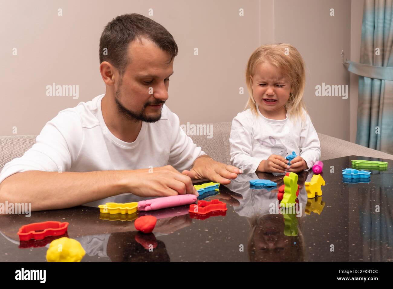 Weinendes kleines Kind und junger kaukasischer Vater spielen mit bunt Teig spielen Stockfoto