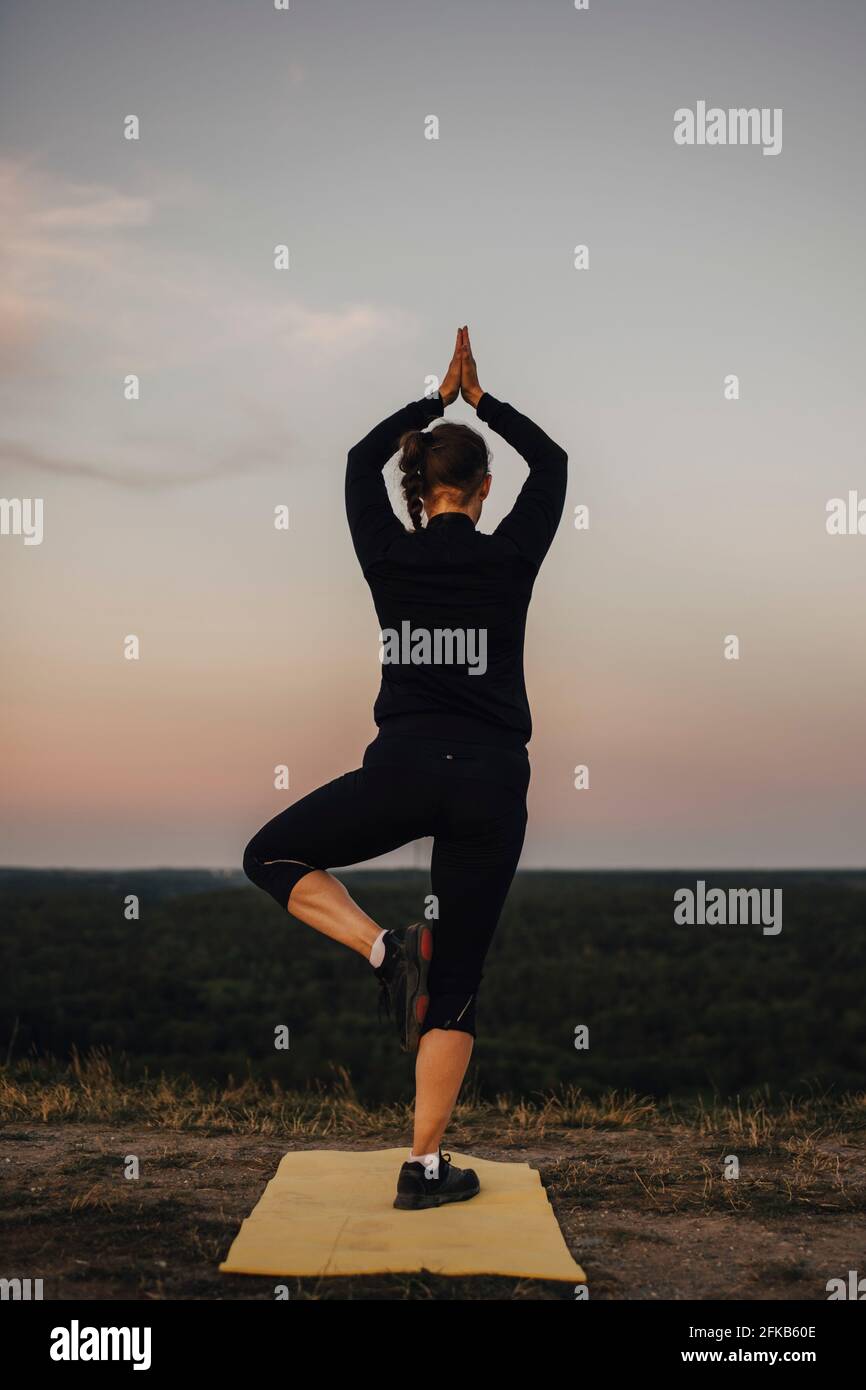 Die reife Sportlerin übt die Baumhaltung bei Sonnenuntergang gegen den Himmel Stockfoto