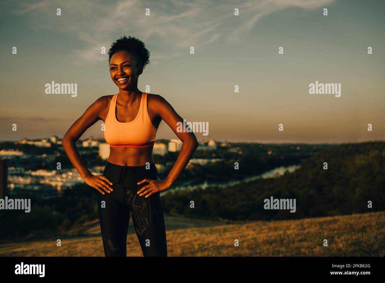 Lächelnde Athletin mit der Hand an der Hüfte während des Sonnenuntergangs Stockfoto