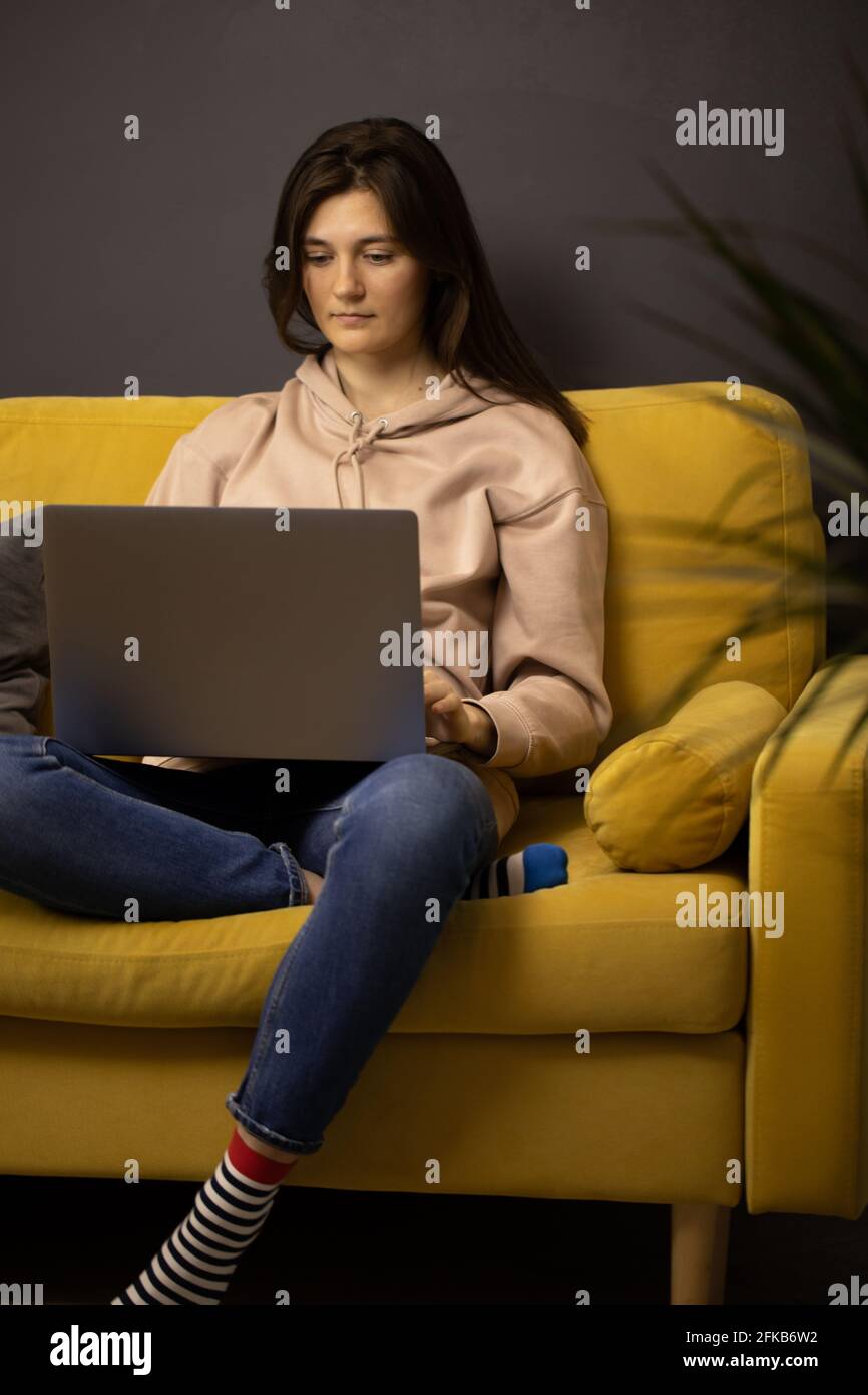Verärgert langhaarige Brünette Mädchen sitzend Knie auf gelbem Sofa gebogen SMS an Laptop senden Stockfoto