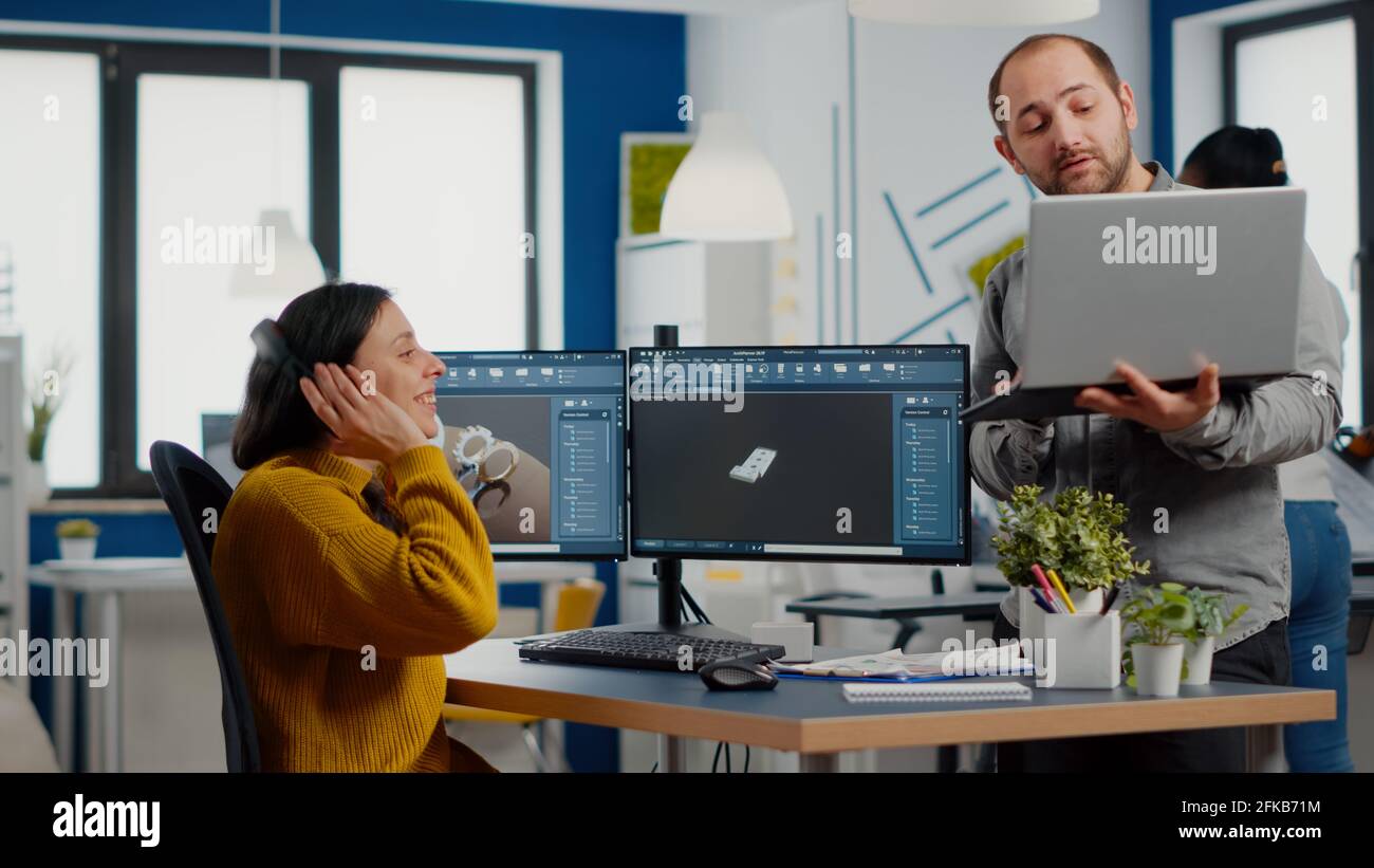 Industriedesignerin diskutiert mit Ingenieurin während der Arbeit in CAD-Programm, Entwicklung neuer Komponenten, Vergleich 3D-Prototyp mit Projekt von Laptop. Arbeitsteam, das Computer mit zwei Monitoren verwendet Stockfoto
