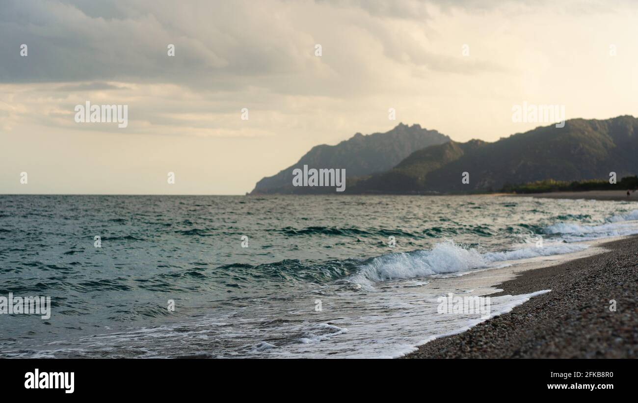 Am Abend Wellen am Strand und in den Bergen. Fokus auf Vordergrund, verschwommener Hintergrund mit Küstenlinie. Stockfoto