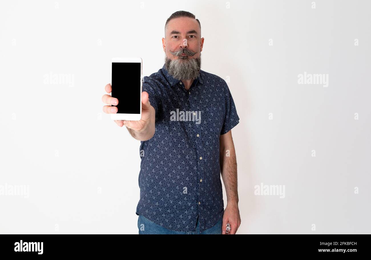 Der kaukasische bärtige Hipster, 40-45 Jahre alt, blickt geradeaus und zeigt den Bildschirm seines Smartphones mit ernstem Gesicht Stockfoto