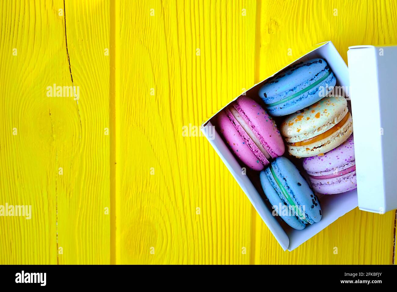 Französische Makronen-Süßigkeiten, verschiedene Geschmacksrichtungen und Farben. Box mit Makronen auf gelbem Holz Holzstruktur Tisch Hintergrund. Geburtstag valentinstag Frühlingsgeschenk Stockfoto