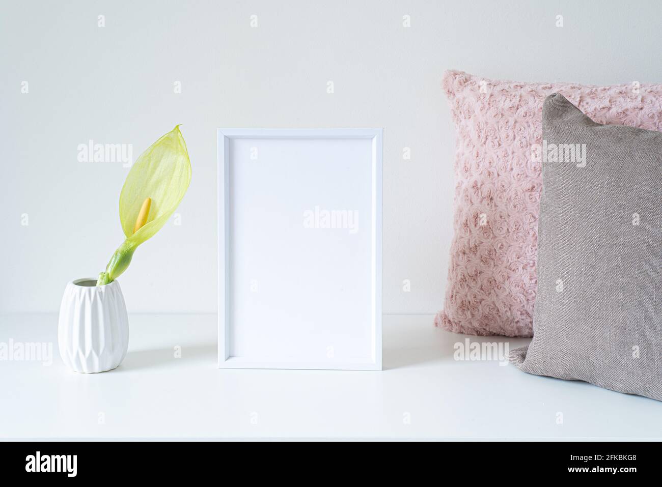 Leeres Modell mit weißem Rahmen auf weißem Tisch. Weiche farbige Kissen und weiße Vase mit einer Arum italicum-Blume. Moderner und minimalistischer Lebensstil. Skandinavisch Stockfoto