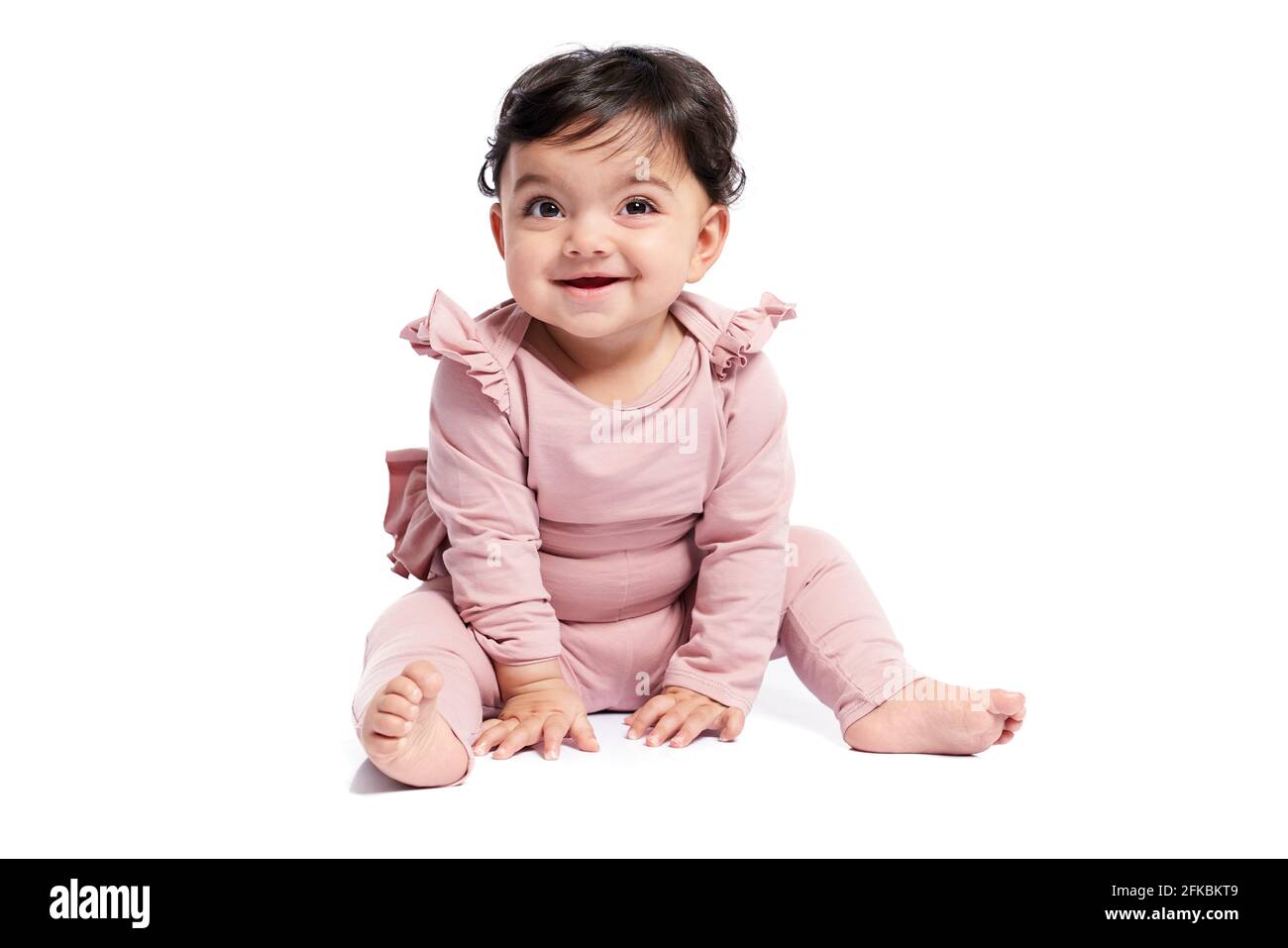 Niedliches weibliches Baby im schönen rosa Outfit lächelnd mit offenem Mund. Attraktives kleines Baby, das auf dem Boden sitzt und posiert, isoliert auf weißem Studiohinterzimmer Stockfoto