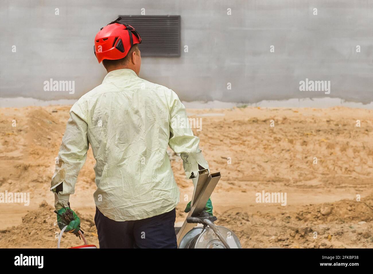 Industriearbeiter in einem roten Helm geht mit einem Arbeitsgerät vor den Hintergrund eines Sand- und Betonbauhauses auf einer Baustelle. Stockfoto
