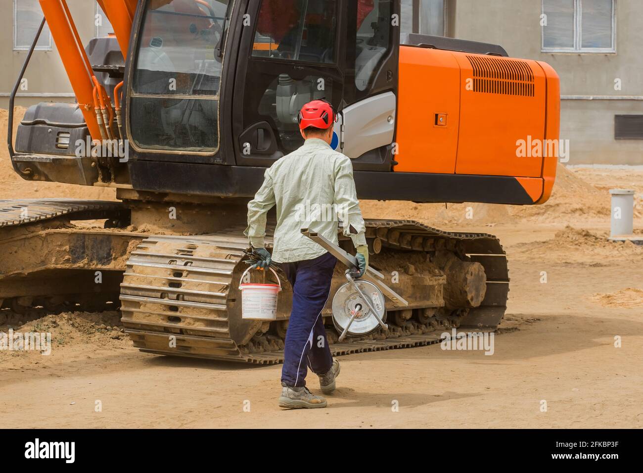 Industriearbeiter in einem roten Helm geht mit einem Arbeitsgerät vor den Hintergrund eines Raupenbaggers auf einer Baustelle. Stockfoto