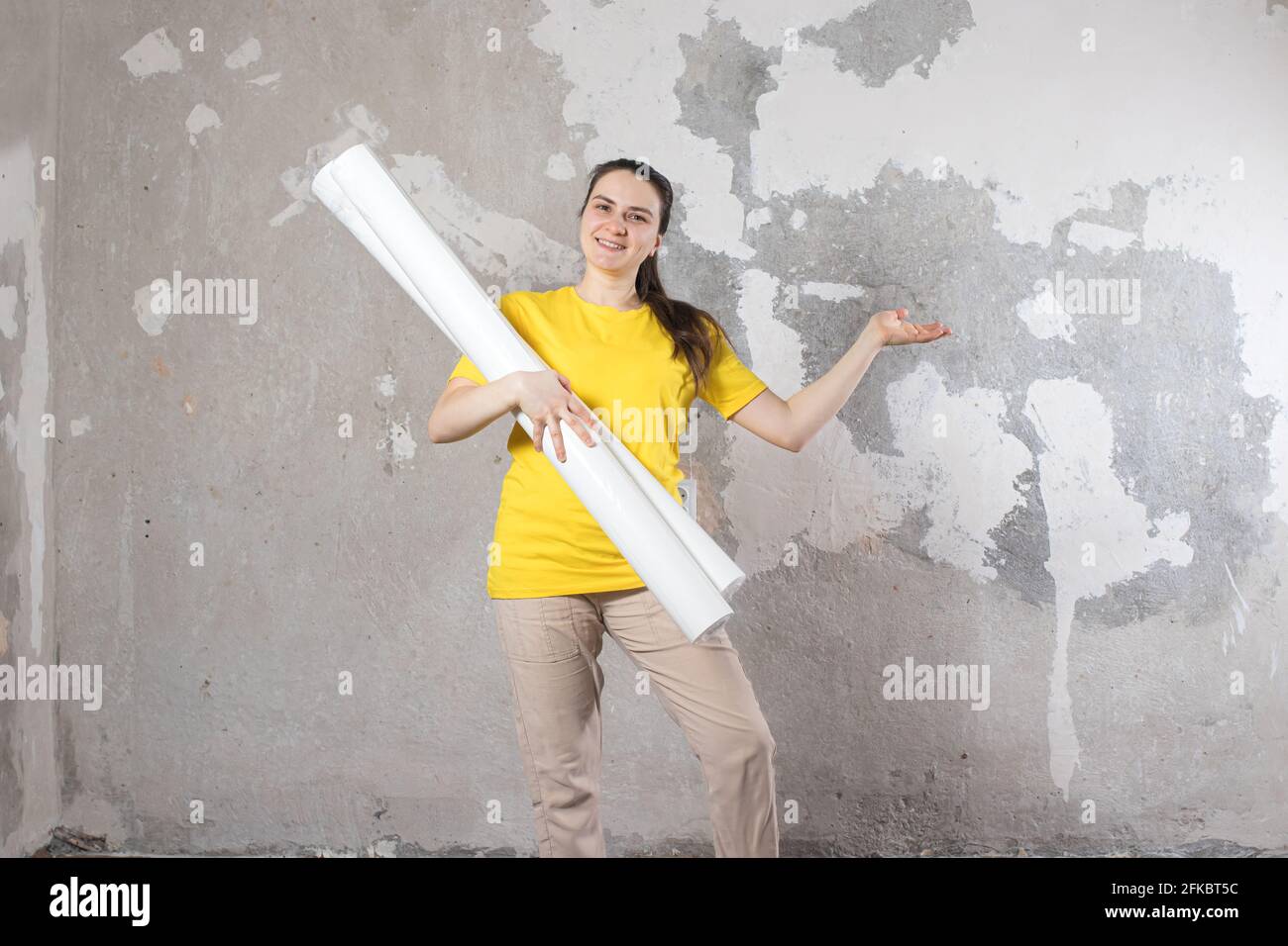 Eine Frau in einem leuchtend gelben T-Shirt hält 2 Rollen weißer Tapete gegen die alte Wand. Reparaturarbeiten, Tapezieren. Mit einem Platz für Text zum Kopieren von SPA Stockfoto