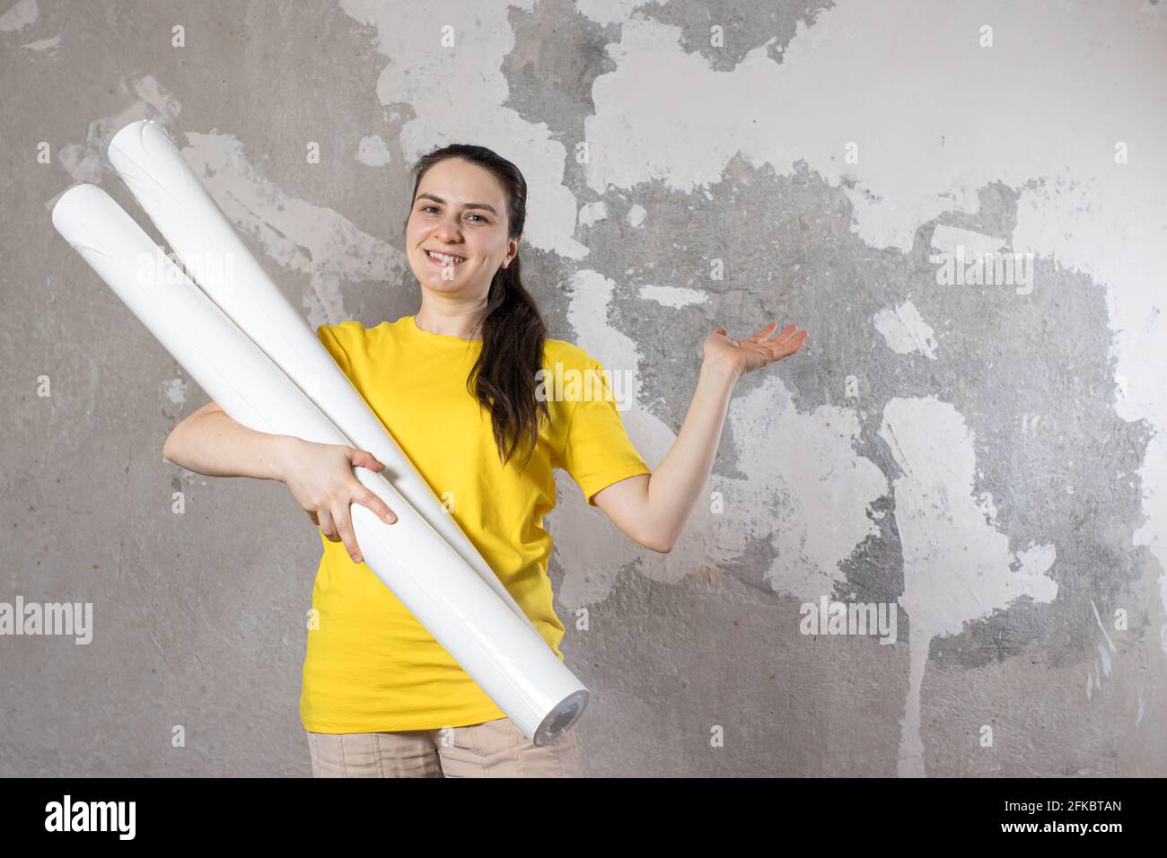 Eine Frau in einem leuchtend gelben T-Shirt hält 2 Rollen weißer Tapete gegen die alte Wand. Reparaturarbeiten, Tapezieren. Mit einem Platz für Text zum Kopieren von SPA Stockfoto