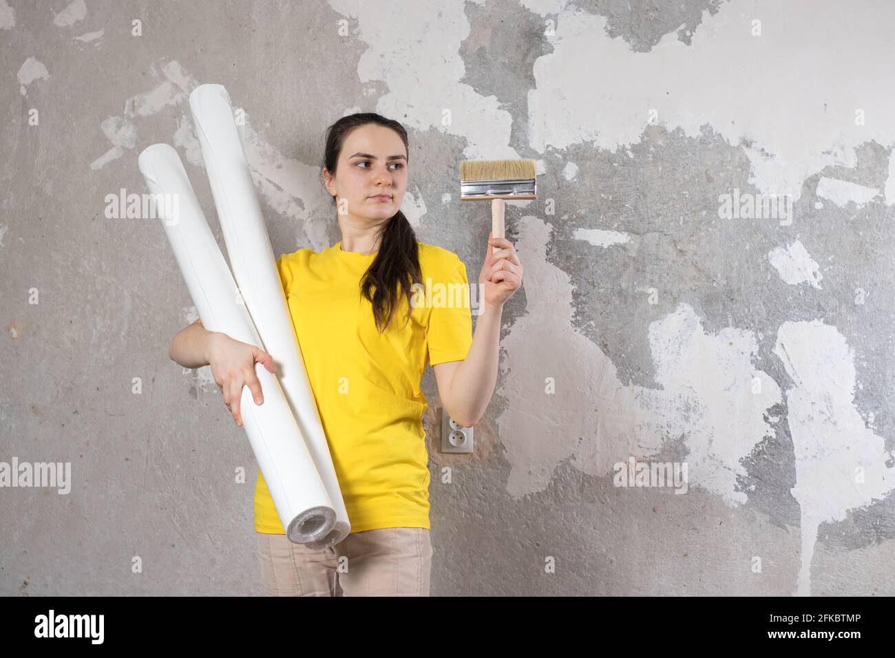 Eine Frau in einem leuchtend gelben T-Shirt hält 2 Rollen weißer Tapete gegen die alte Wand. Reparaturarbeiten, Tapezieren. Mit einem Platz für Text zum Kopieren von SPA Stockfoto