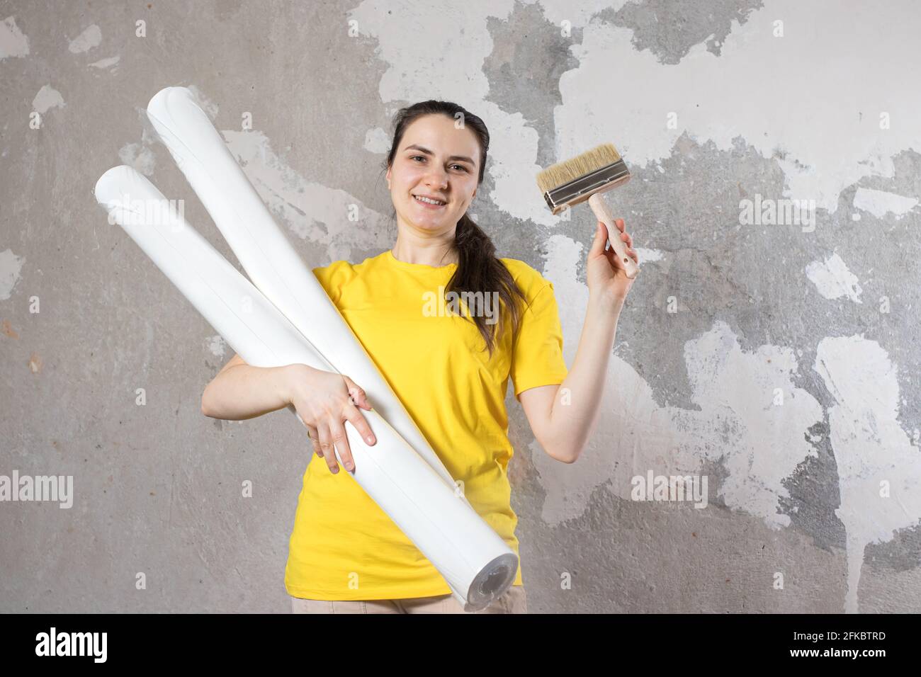 Eine Frau in einem leuchtend gelben T-Shirt hält 2 Rollen weißer Tapete gegen die alte Wand. Reparaturarbeiten, Tapezieren. Mit einem Platz für Text zum Kopieren von SPA Stockfoto