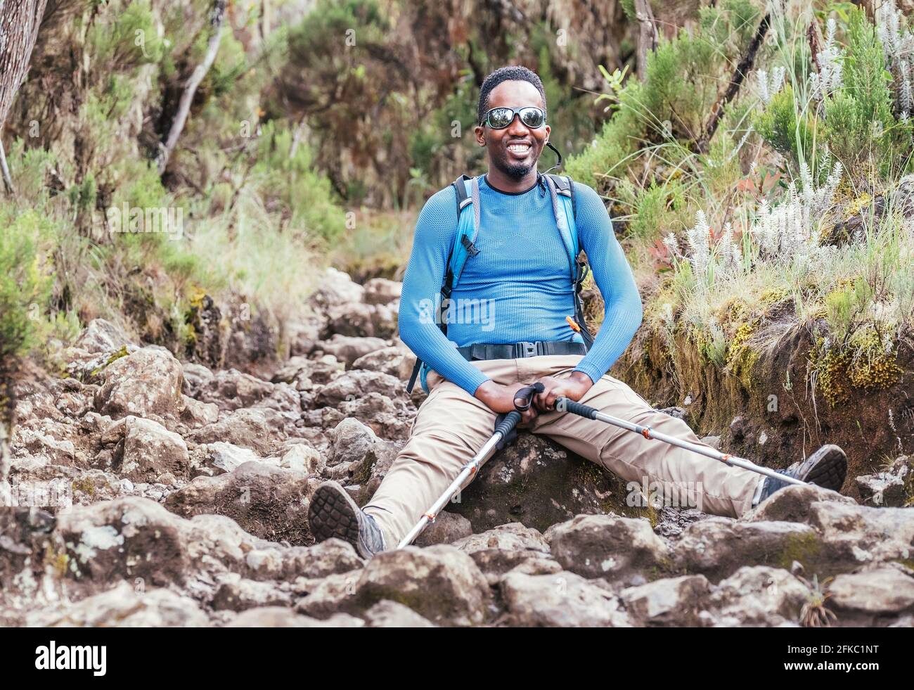 Porträt eines fröhlich lächelnden afroamerikanischen Ethnizität jungen Mannes in Sonnenbrillen sitzen mit Rucksack und Trekkingstöcke und ruhen auf Berg t Stockfoto