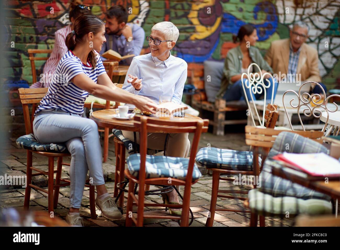 Eine ältere Frau und ihre junge Freundin sprechen über ein Buch, während sie in entspannter Atmosphäre in der Bar einen Drink genießen. Freizeit, Bar, Freundschaft, Stockfoto