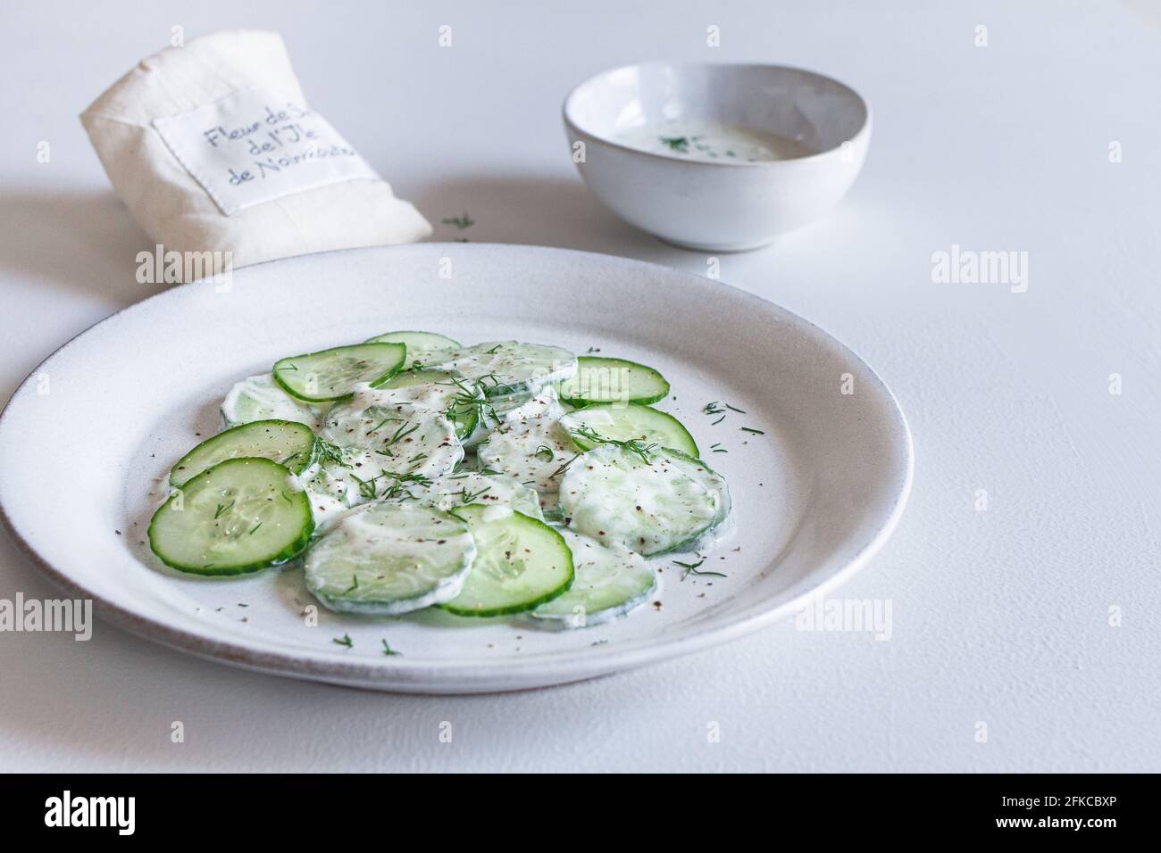 Teller mit cremigem Joghurt-Gurkensalat mit einer Schüssel Joghurt-Dressing und einer Tüte Fleur de Sel im Hintergrund. Stockfoto