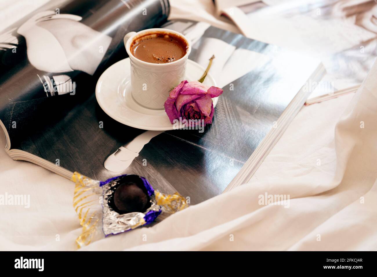 Selektive Fokus EIN türkischer Kaffeebecher und eine getrocknete Rose auf dem Teller davon . Einige öffnen Zeitschriften darunter und eine Schokolade bereit zu beißen. Stockfoto